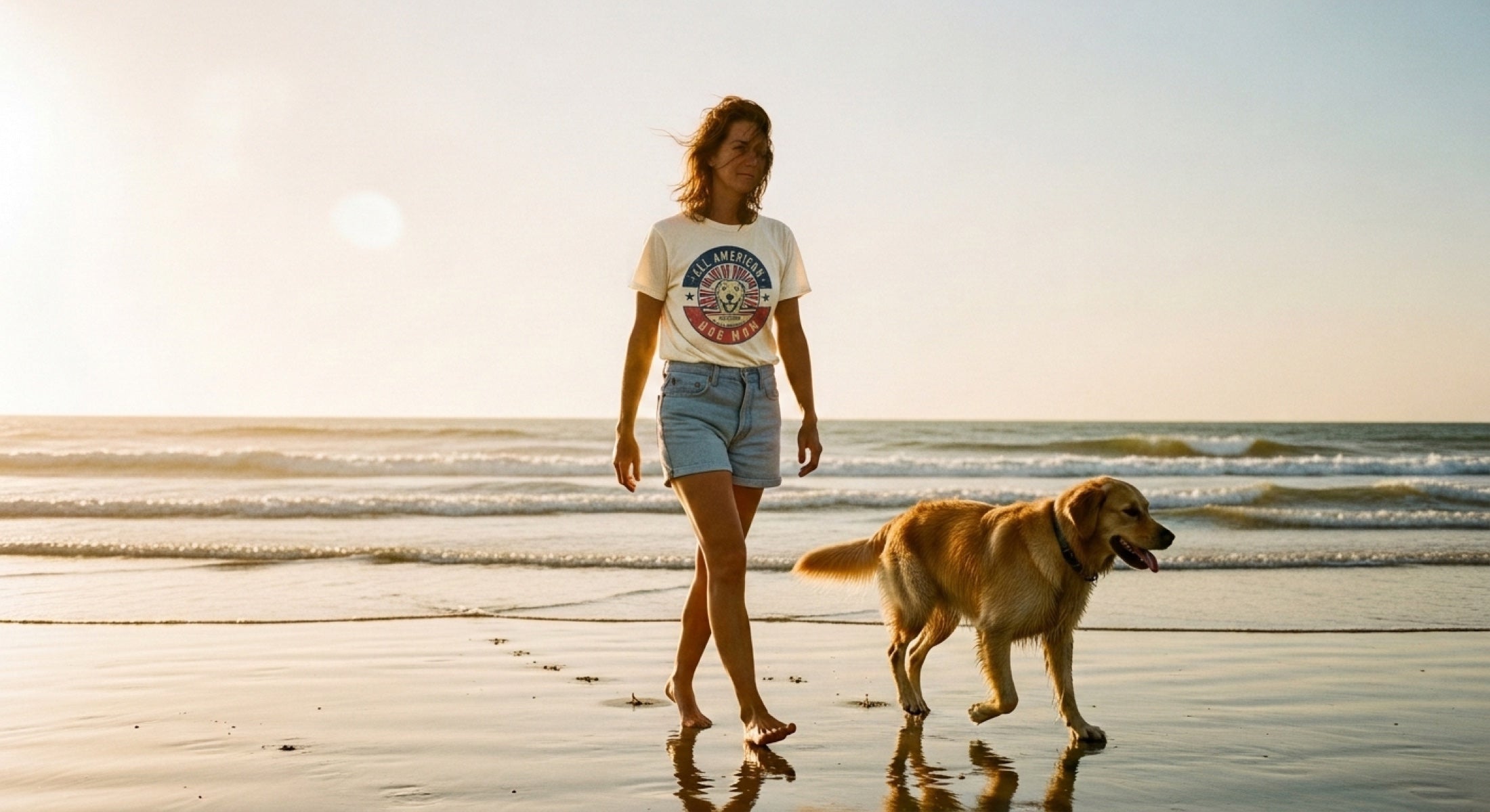 Woman in an All American Dog Mom tee walking her golden retriever on a Carolina beach at golden hour